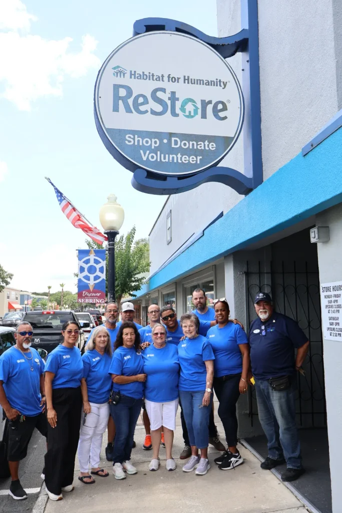 Habitat ReStore Sebring volunteers and staff in blue shirts standing beside the round Habitat ReStore sign on the storefront